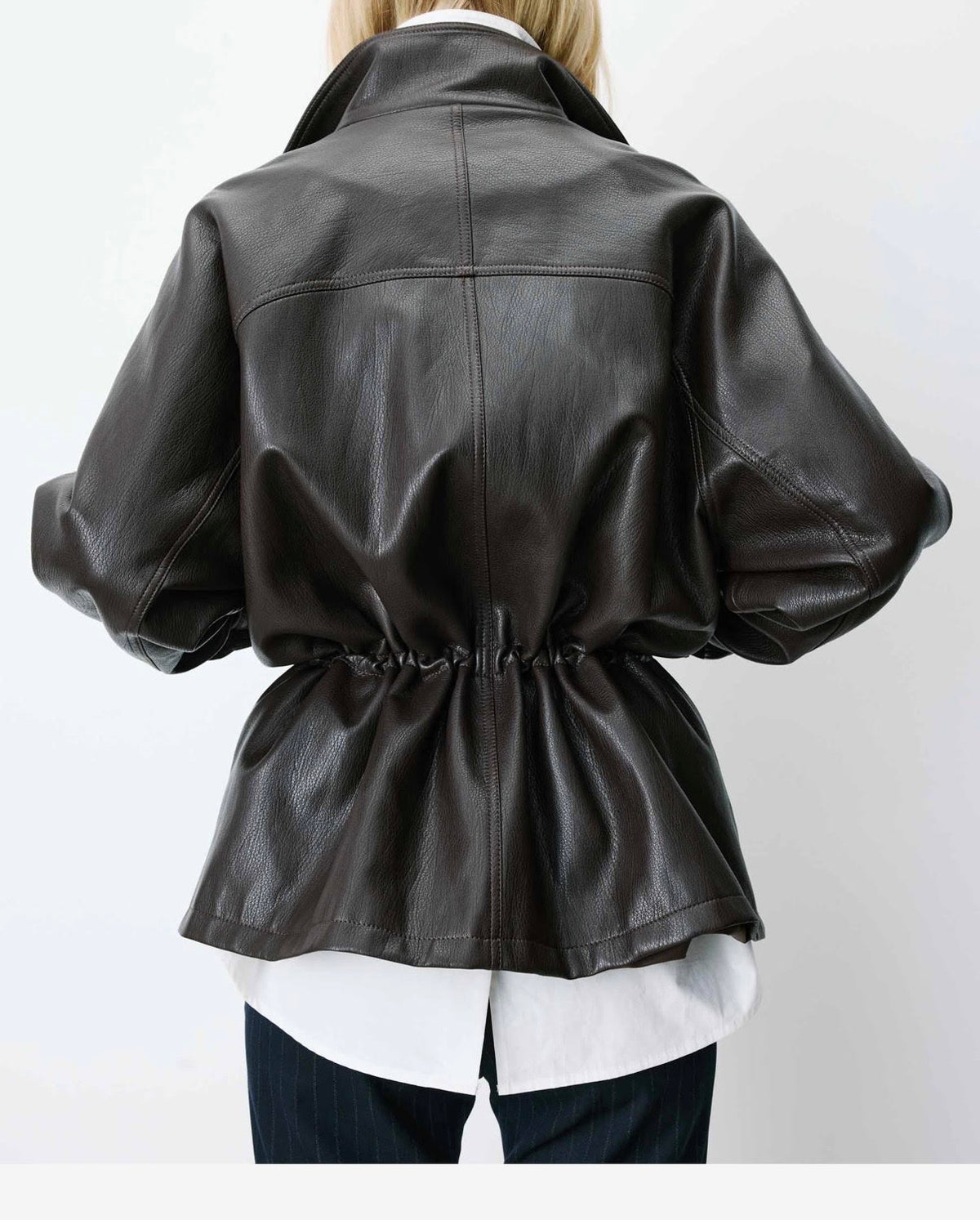 A person shown from the back wearing an oversized dark brown faux-leather jacket with a gathered waist, layered over a white shirt and dark pants, against a plain white background.