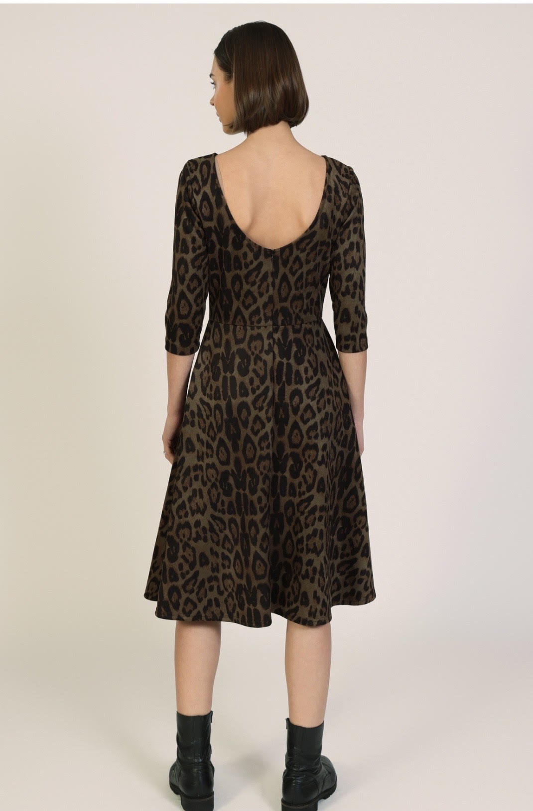 A woman viewed from the back wearing a brown and black leopard-print dress with three-quarter sleeves and a deep open back, paired with black ankle boots, against a plain light beige studio background.