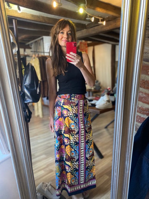 A woman takes a mirror selfie in a boutique, wearing a black sleeveless top and a long, colorful floral print skirt with a patterned border. The boutique has wooden floors, brick walls, and hanging clothing in the background.