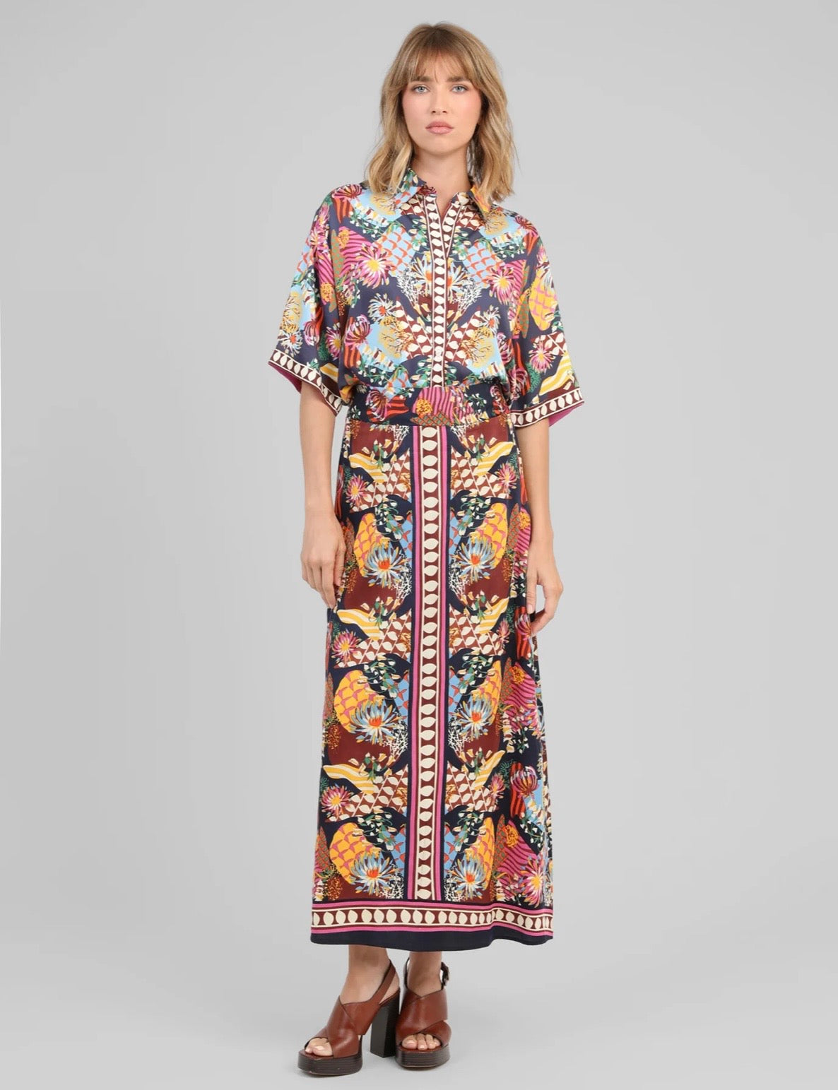 A woman modeling a long-sleeved, button-front maxi dress with a vibrant multicolor abstract pattern and a matching self-tie belt, paired with brown platform sandals, against a solid gray studio background.