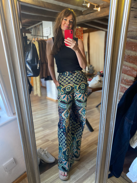A woman takes a mirror selfie in a boutique, wearing high-waisted wide-leg pants with a colorful geometric medallion pattern in teal, gold, and black, paired with a black sleeveless top.