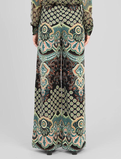 A person shown from the back wearing wide-leg trousers with an ornate multicolor paisley and geometric print in black, teal, beige, and rust tones, against a light gray studio background.