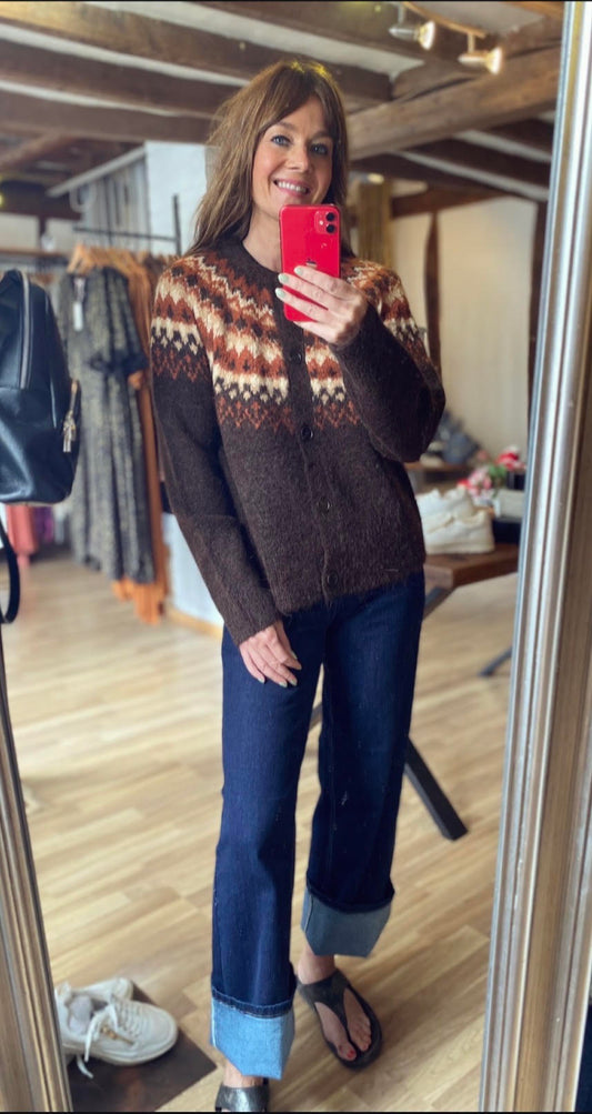 A woman takes a mirror selfie in a clothing store, wearing a brown knit cardigan with a colorful geometric pattern across the chest and dark blue jeans.
