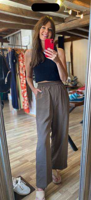 A woman takes a mirror selfie in a clothing boutique, wearing a dark navy sleeveless top and high-waisted, wide-leg taupe-brown pants. The boutique has wooden floors, clothing racks, and warm lighting.