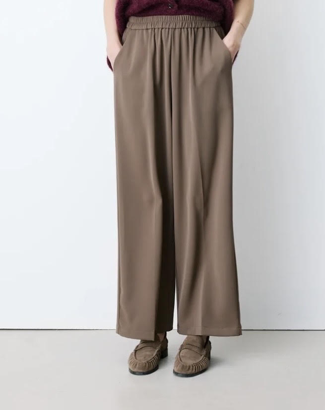 A person wearing loose, wide-leg taupe-brown pants with side pockets, paired with brown suede moccasin-style shoes, shown from the waist down against a plain light gray studio background.