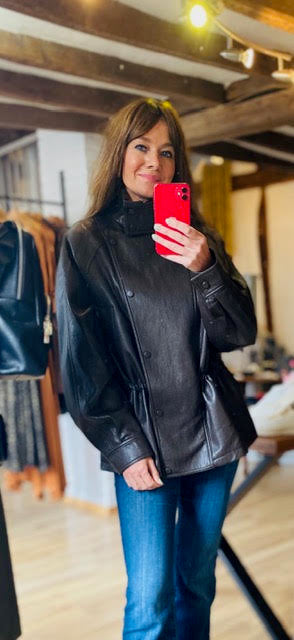 A woman takes a mirror selfie in a clothing boutique wearing a black leather jacket, blue jeans, and a black turtleneck, holding a red smartphone.