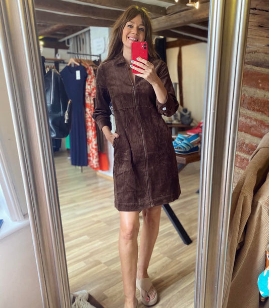 A woman takes a mirror selfie in a boutique while wearing a dark brown corduroy zip-front mini coat. The boutique interior features wood floors, clothing racks, and a brick wall in the background.