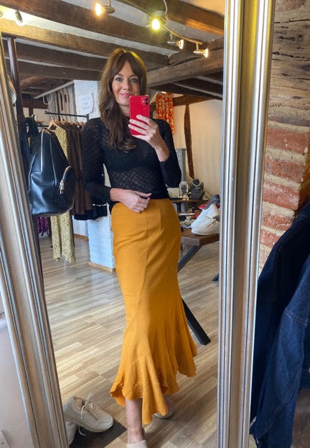 A woman takes a mirror selfie in a boutique, wearing a black long-sleeve mesh top and a mustard yellow flared midi skirt. The boutique has wooden floors, brick walls, and clothing racks in the background.