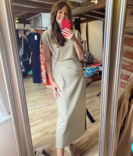 A woman takes a mirror selfie in a boutique while wearing a short-sleeved, knee-length beige dress with a twisted detail at the waist. The boutique has wooden floors, brick walls, and clothing racks in the background.