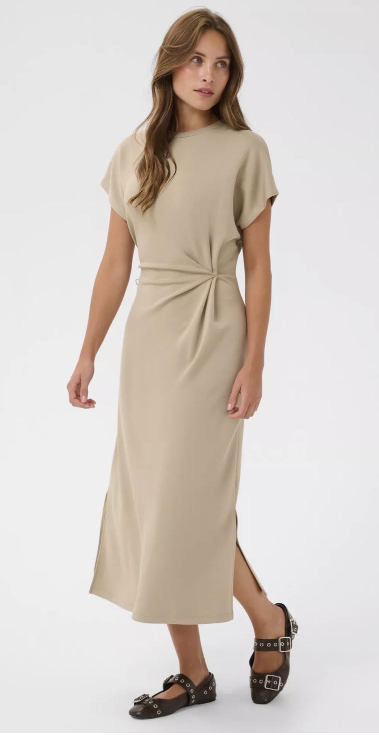 A woman modeling a beige short-sleeve midi dress with a twisted waist detail, paired with black strappy sandals, against a plain white studio background.