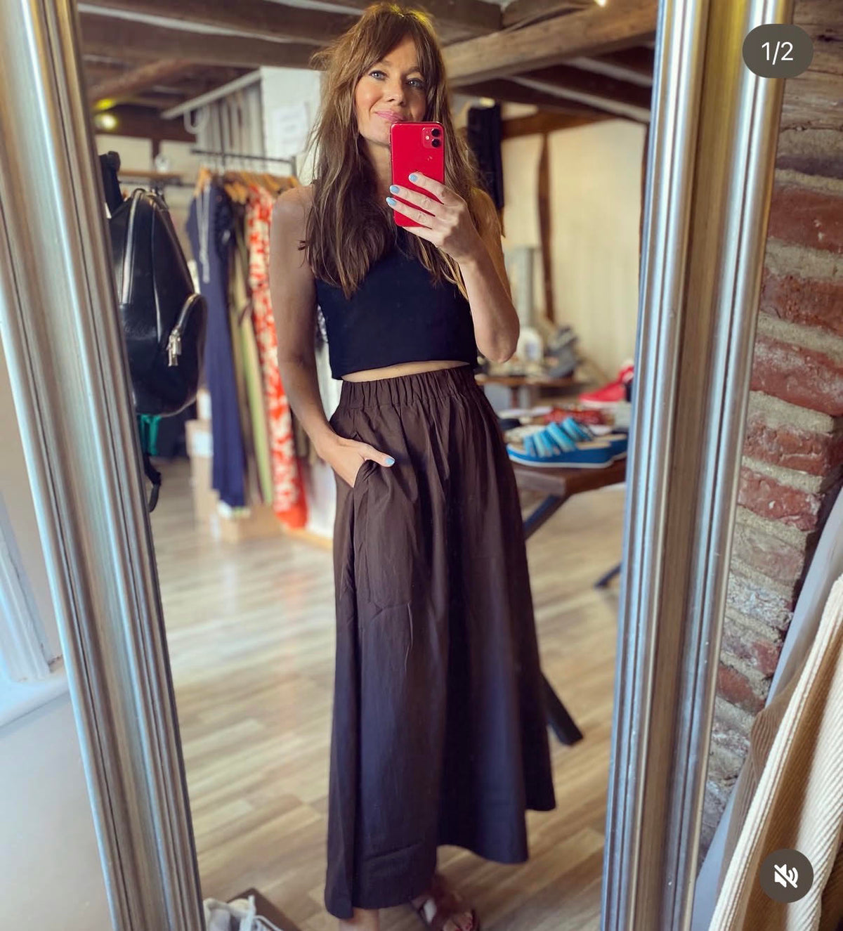A woman takes a mirror selfie in a boutique, wearing a black halter crop top and a dark brown high-waisted maxi skirt. The boutique has wooden floors, brick walls, and clothing racks in the background.