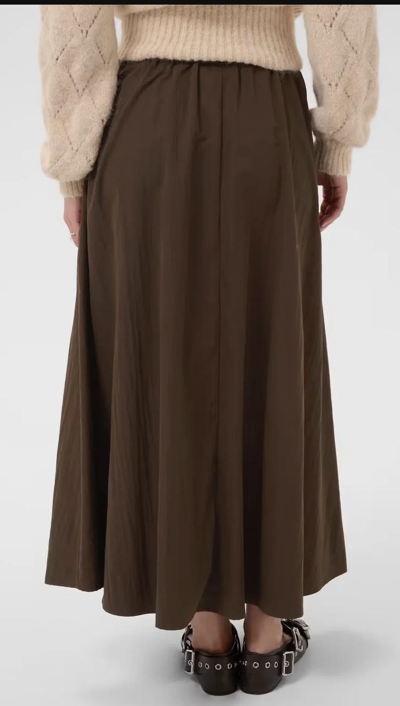 A person shown from the back wearing a long, pleated brown skirt, a cream-colored knit sweater, and dark brown studded shoes against a plain light gray studio background.