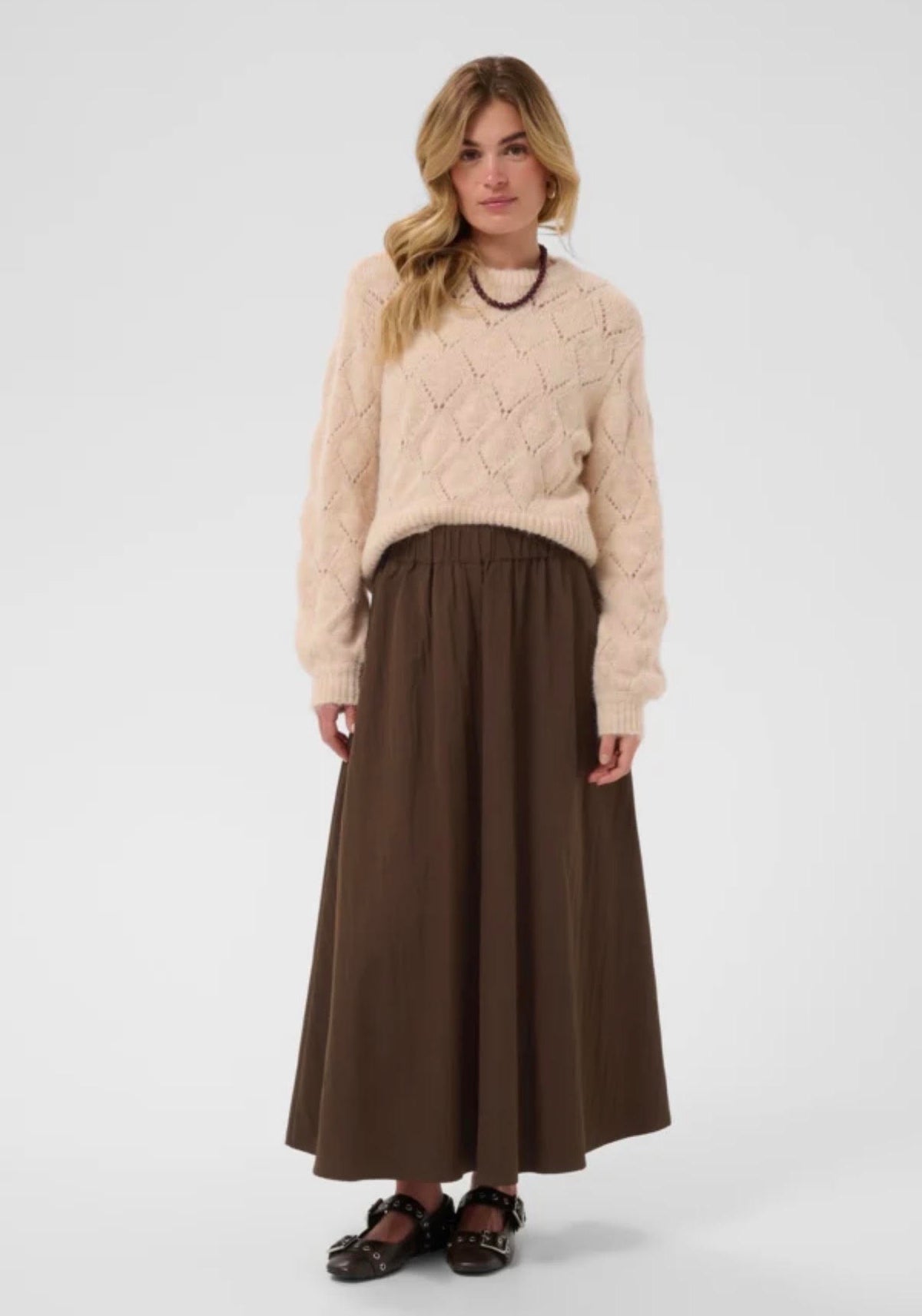 A woman modeling a cream cable-knit sweater and a brown pleated maxi skirt against a light gray studio background.