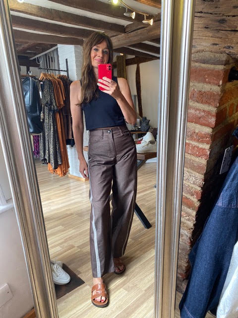 A woman takes a mirror selfie in a clothing boutique, wearing brown wide-leg cropped trousers with white side stripes, a black sleeveless top, and brown sandals.