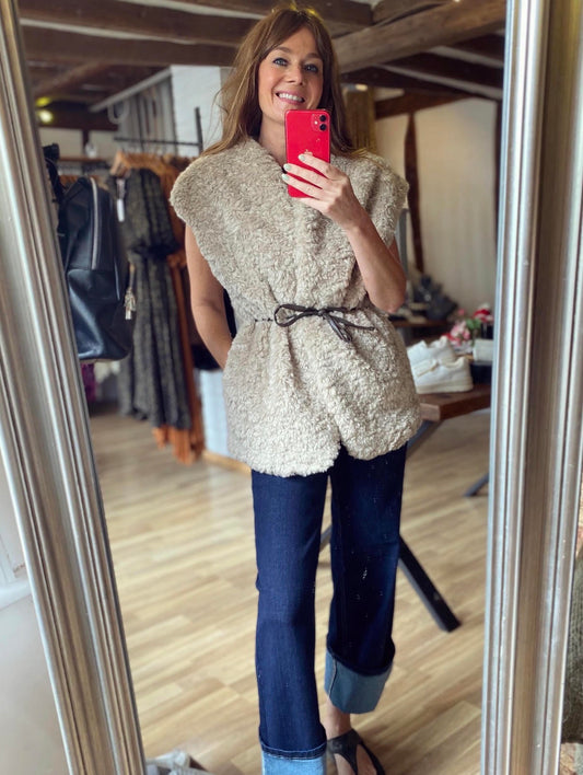A woman takes a mirror selfie in a clothing boutique, wearing a beige fuzzy sleeveless gilet with a black tie belt, dark blue jeans, and black sandals.