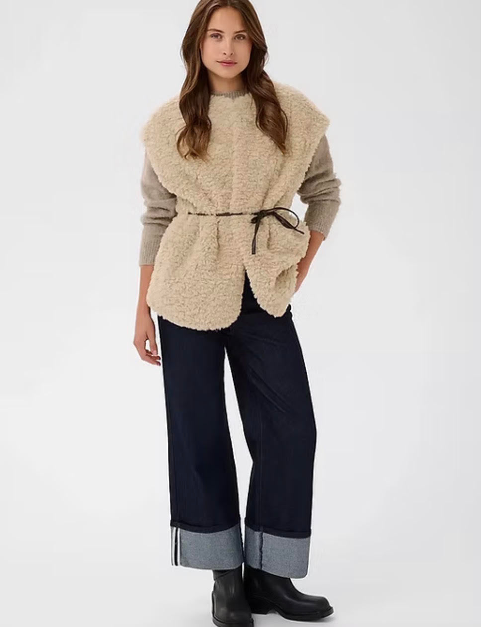 A woman modeling a beige sherpa fleece vest with a tie waist, worn over a taupe long-sleeve top and dark wide-leg jeans with cuffed hems, against a plain light gray studio background.