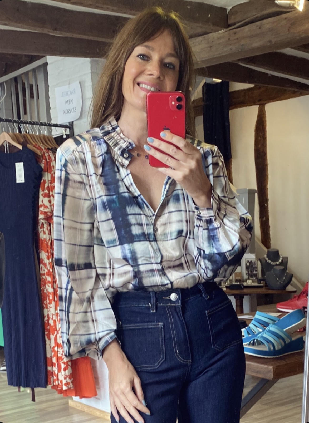 A woman takes a mirror selfie in a boutique, wearing a blue, white, and beige plaid button-down shirt tucked into dark wash jeans. She holds a red smartphone to capture the photo, with clothing racks and wooden ceiling beams visible in the background.