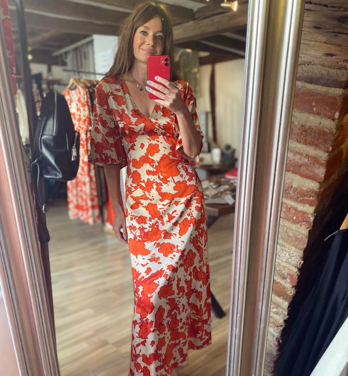 A woman takes a mirror selfie in a boutique wearing a red and orange abstract floral print maxi dress with short flutter sleeves and a deep V-neck. The background shows other clothing racks and a brick wall.