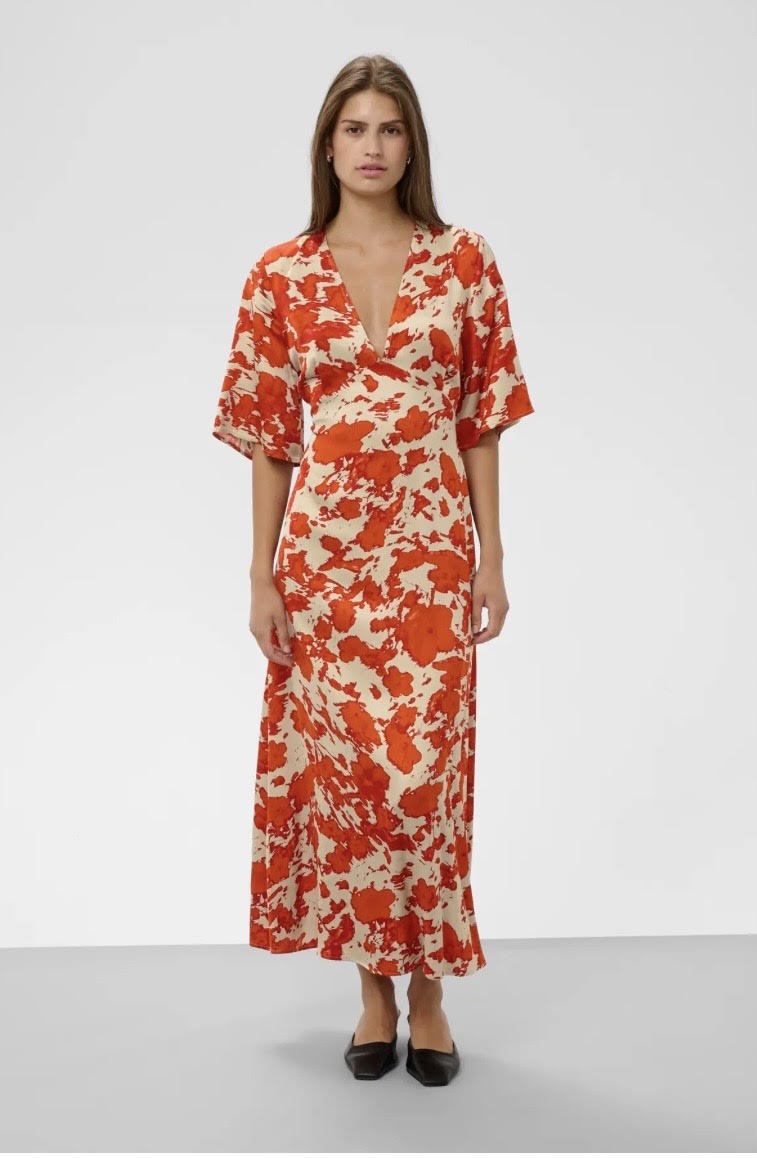 A woman modeling a short-sleeved midi wrap dress with a vibrant orange and cream abstract floral pattern, standing against a light gray studio background.