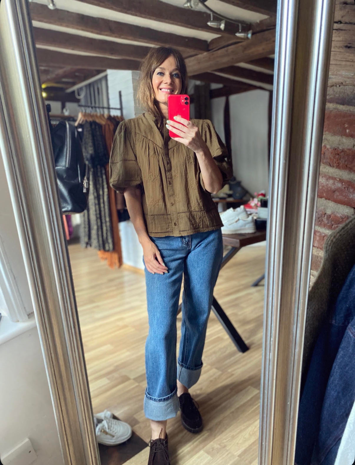 A woman takes a mirror selfie in a boutique, wearing a brown short-sleeve button-up shirt, blue cuffed jeans, and dark brown shoes. The boutique has wooden floors, brick walls, and clothing racks in the background.