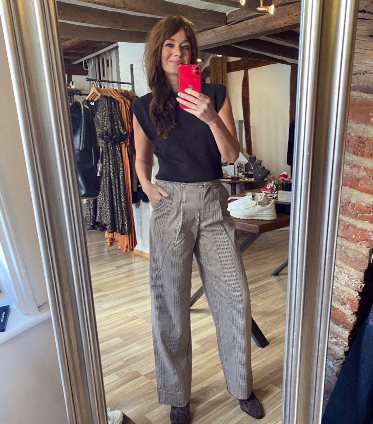 A woman takes a mirror selfie in a boutique, wearing a black sleeveless top and high-waisted, wide-leg gray pinstripe trousers. The boutique has wooden floors, brick walls, and clothing racks in the background.