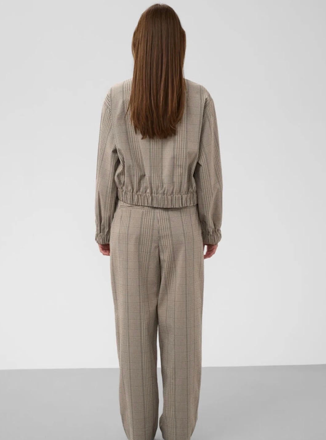 A model viewed from the back wearing a matching beige and brown plaid long-sleeve crop top and wide-leg trousers set, against a plain light gray studio background.