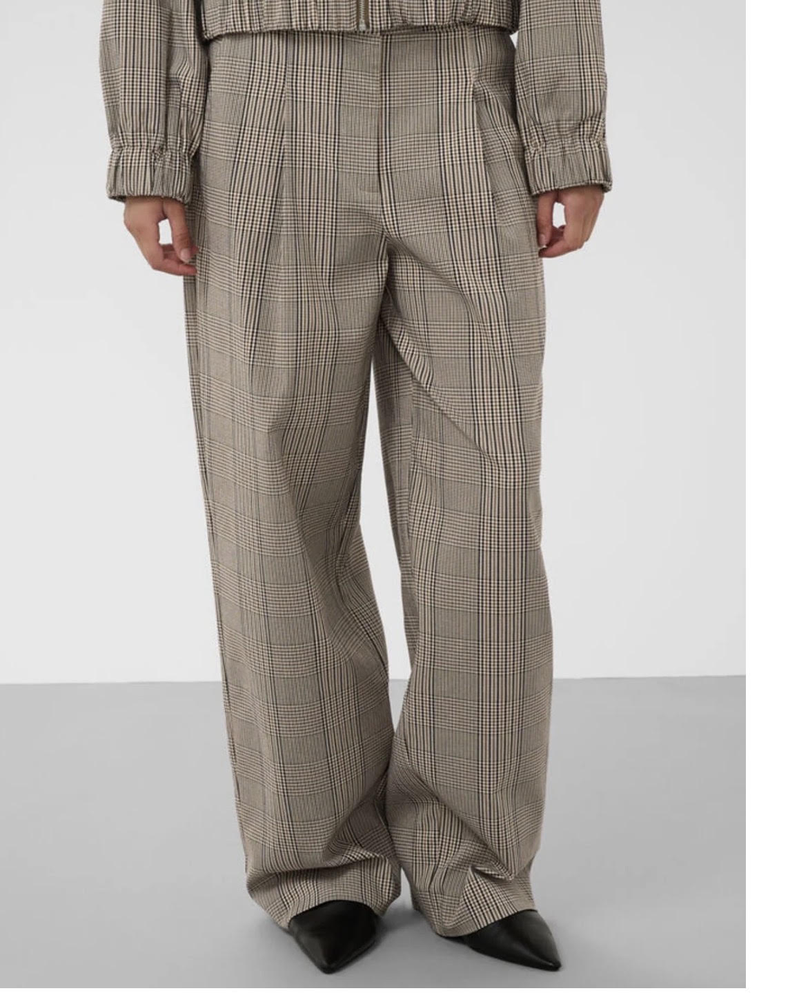 A person wearing wide-leg pleated trousers in a beige and brown plaid pattern, paired with black pointed-toe heels, shown from the waist down against a light gray studio background.
