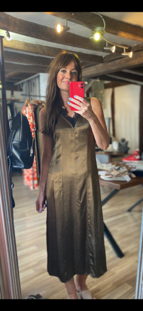 A woman takes a mirror selfie in a boutique while wearing a sleeveless, satin-like olive green midi dress. The boutique has wooden ceiling beams, string lights, and other clothing racks in the background.