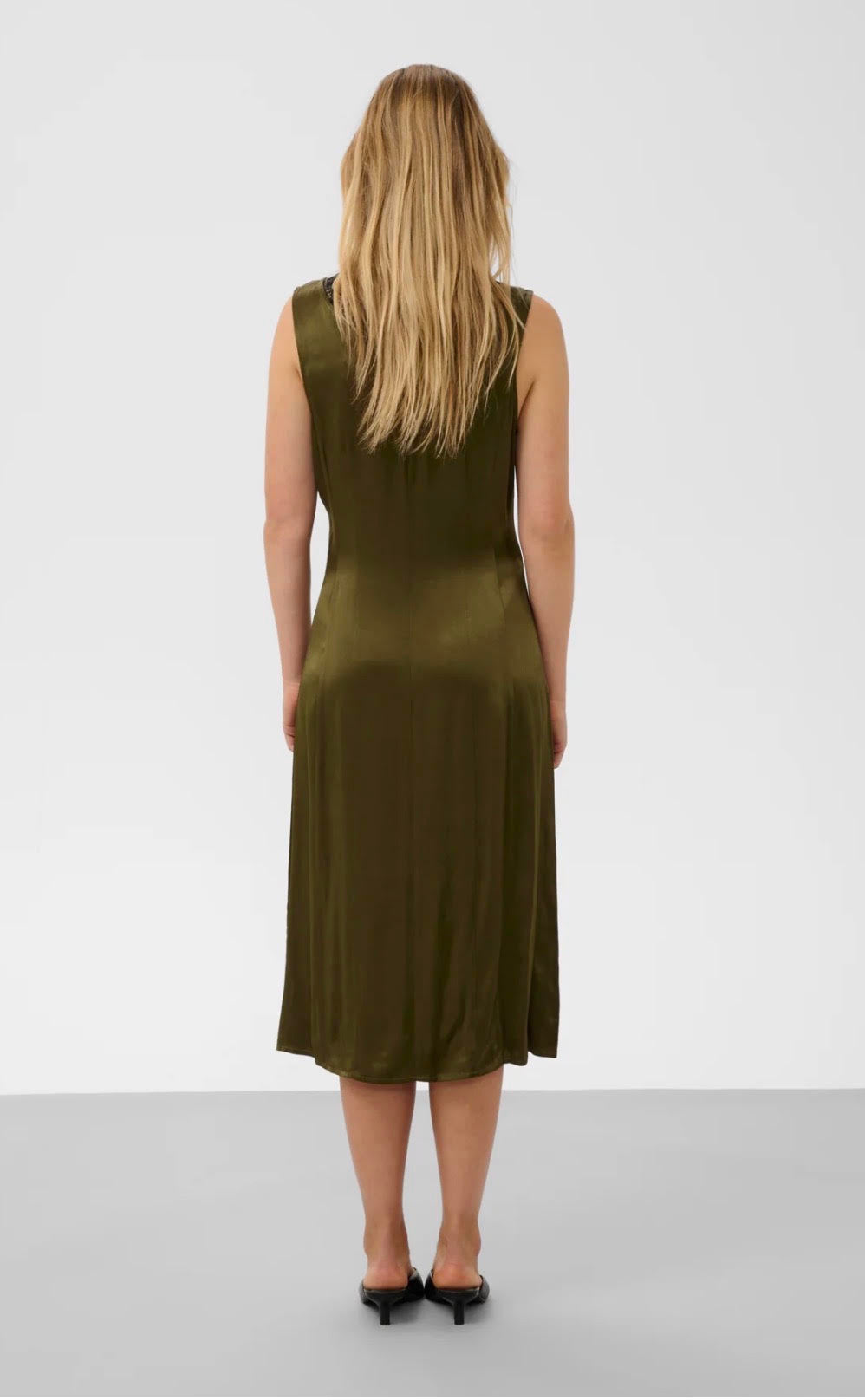 A woman viewed from behind wearing a sleeveless olive green midi dress with a satin-like sheen, paired with black heels, against a light gray studio background.
