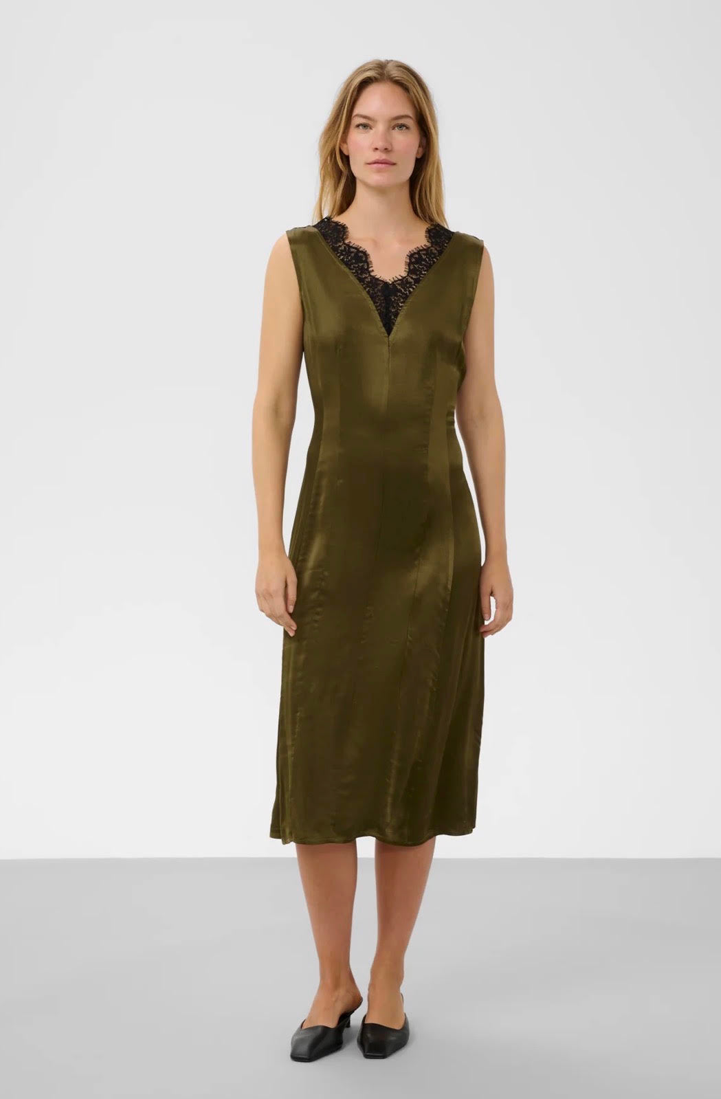A woman modeling a sleeveless olive green midi dress with a black lace V-neckline, standing against a light gray studio background.