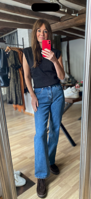 A woman takes a mirror selfie in a boutique, wearing a black sleeveless top, blue flare jeans, and brown lace-up boots. The boutique has wooden floors, clothing racks, and wooden ceiling beams.