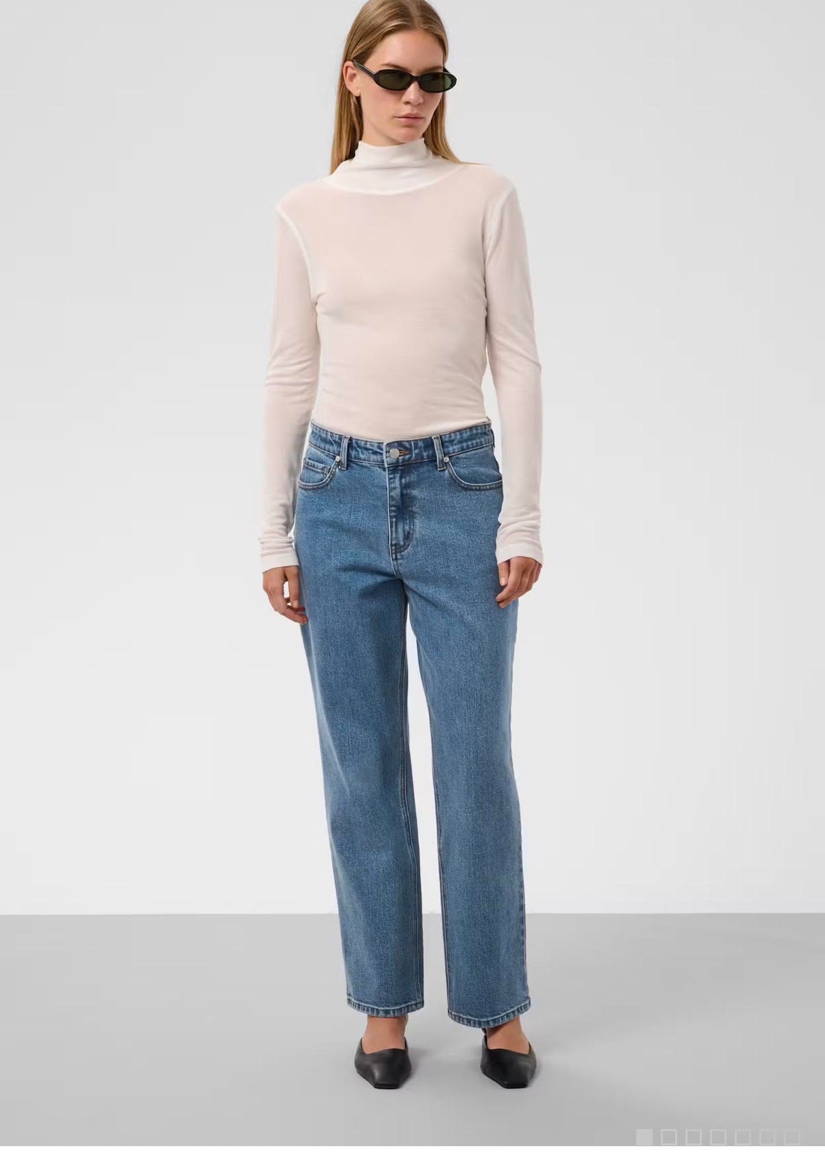A model wearing a pale pink mock-neck long-sleeve top, blue straight-leg jeans, and black pointed flats, standing against a light gray studio background.