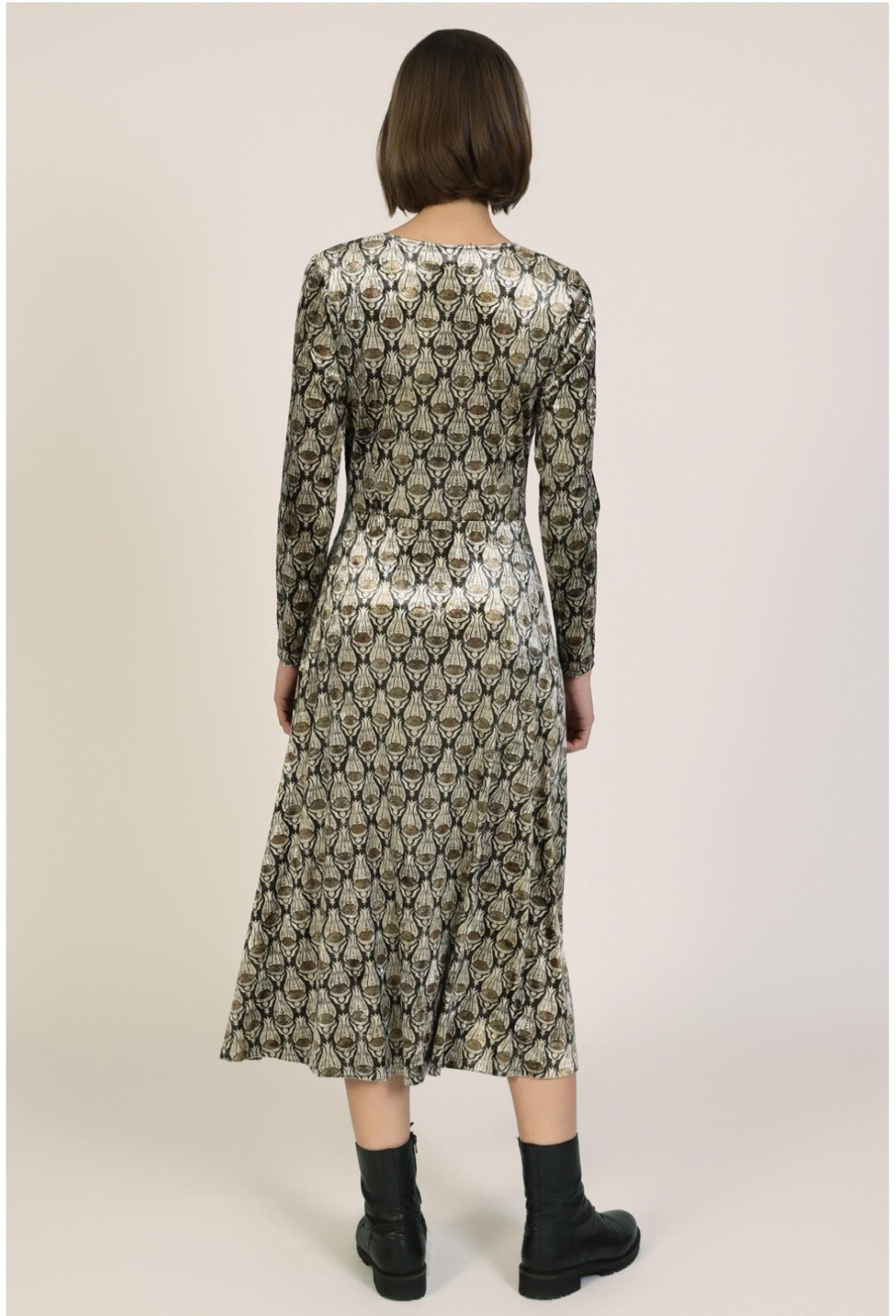 A woman viewed from the back wearing a long-sleeve midi dress with an all-over snakeskin pattern in olive green, beige, and black, paired with black ankle socks and black boots, against a plain light beige studio background.