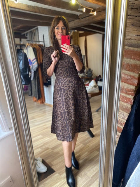A woman takes a mirror selfie in a boutique wearing a brown and black leopard print midi dress with elbow-length sleeves and black ankle boots.