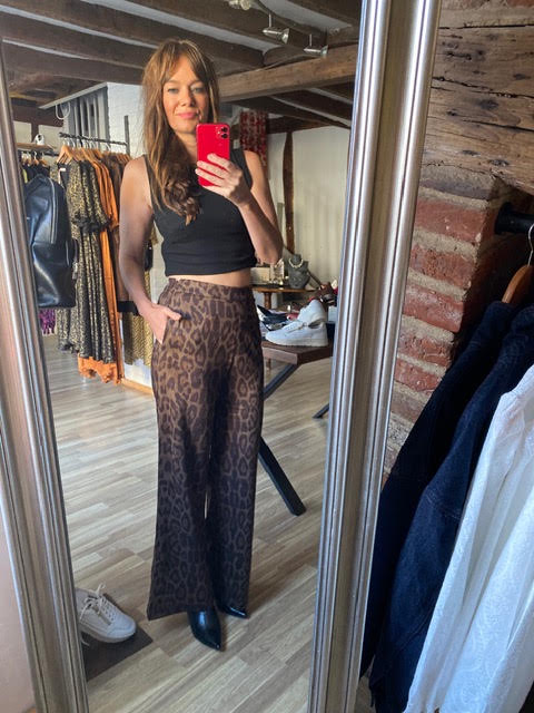 A woman takes a mirror selfie in a boutique, wearing a black sleeveless crop top and brown leopard-print wide-leg pants. The boutique has a rustic interior with exposed wood ceiling beams, brick walls, and clothing racks.