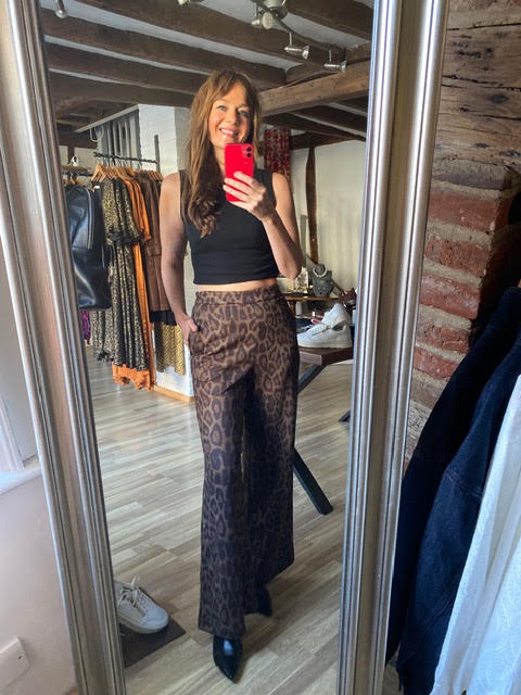 A woman takes a mirror selfie in a boutique, wearing a black crop top and brown leopard-print wide-leg pants. The boutique has wooden floors, brick walls, and clothing racks in the background.