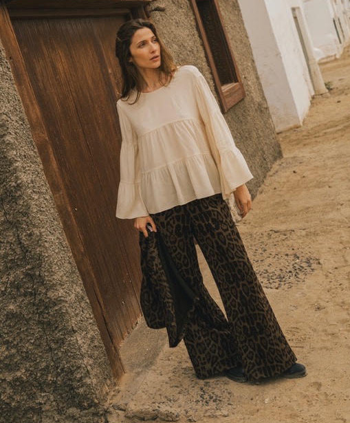 A woman stands outdoors in a narrow street beside a rustic wooden door and textured wall, wearing a cream peplum blouse with dramatic flared bell sleeves and wide-leg black leopard-print pants.