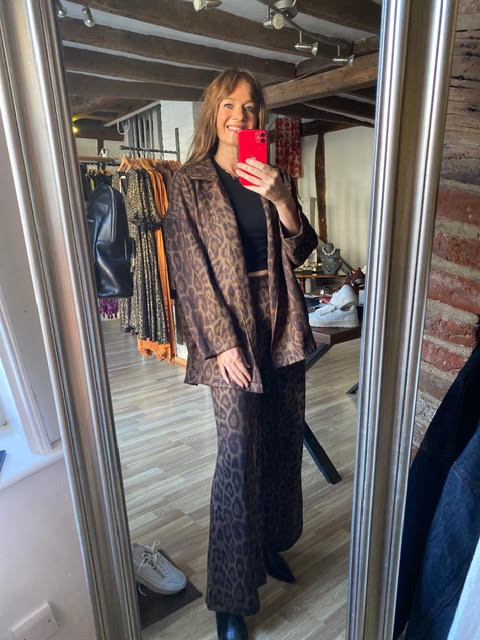 A woman takes a mirror selfie in a clothing boutique wearing a brown and black leopard print blazer and matching wide-leg trousers over a black top.