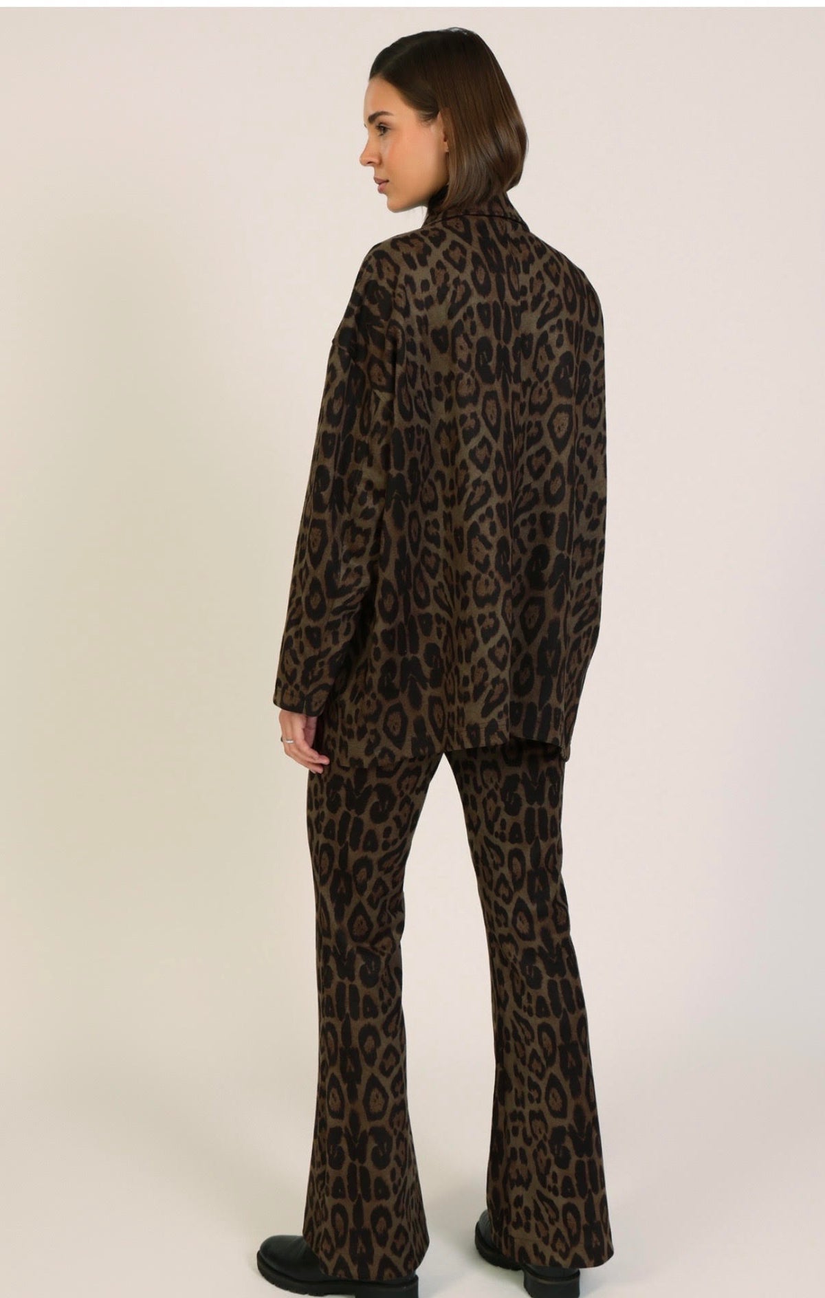 A woman shown from the back wearing a matching brown and black leopard-print blazer and flared pants set, photographed against a plain light beige studio background.