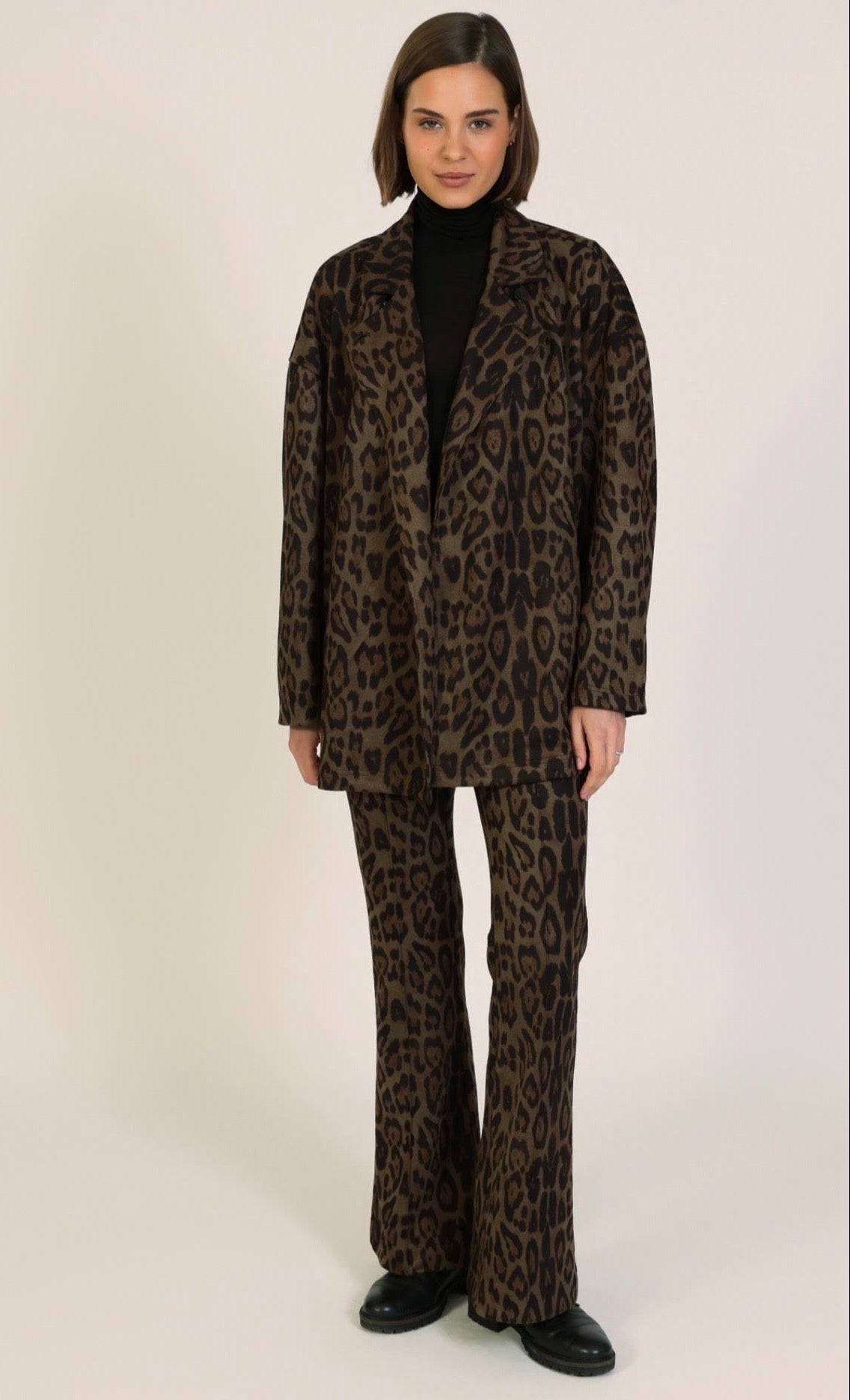A model wearing a matching brown and black leopard-print blazer and wide-leg trousers suit, styled with a black turtleneck and black leather shoes, against a plain light beige studio background.