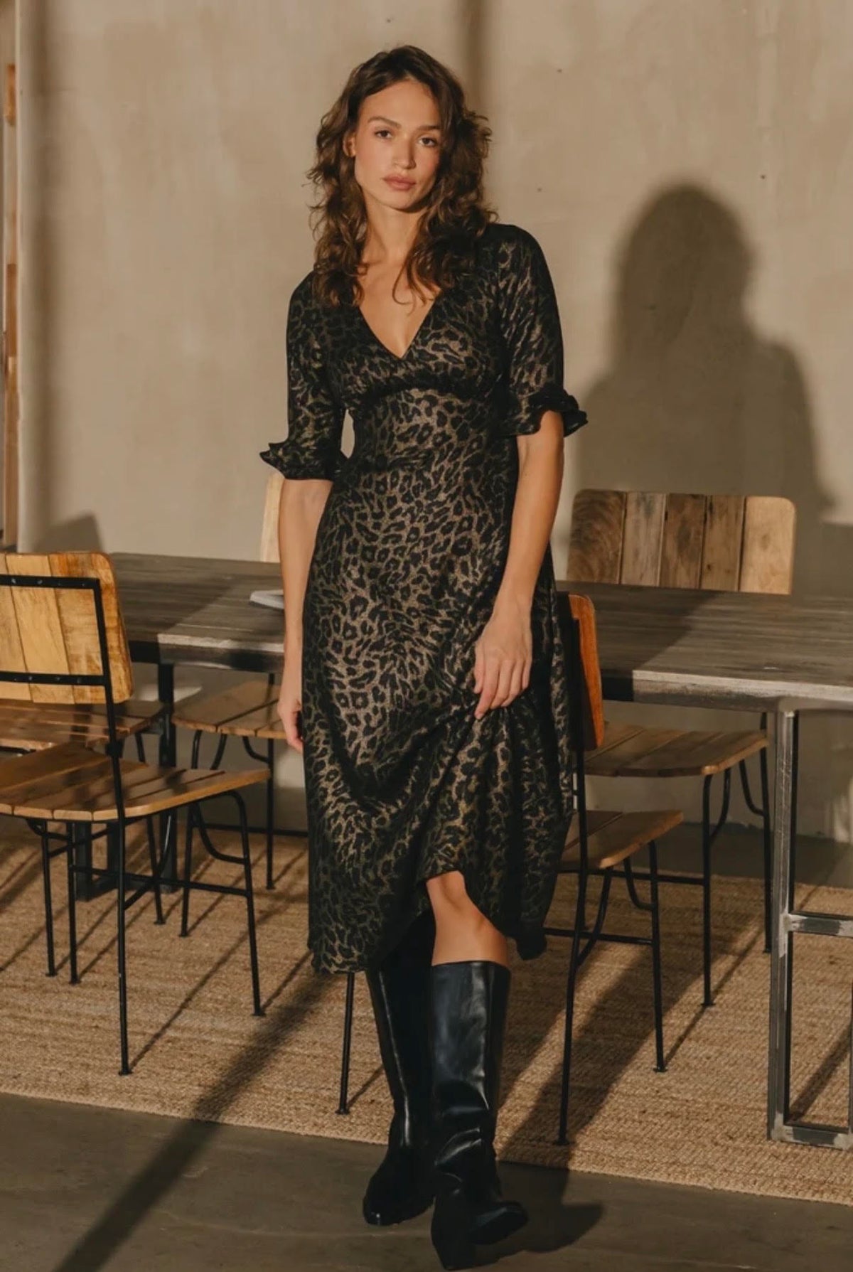 A woman stands in a rustic indoor setting wearing a black leopard-print midi dress with short puff sleeves and a V-neckline, paired with tall black boots.