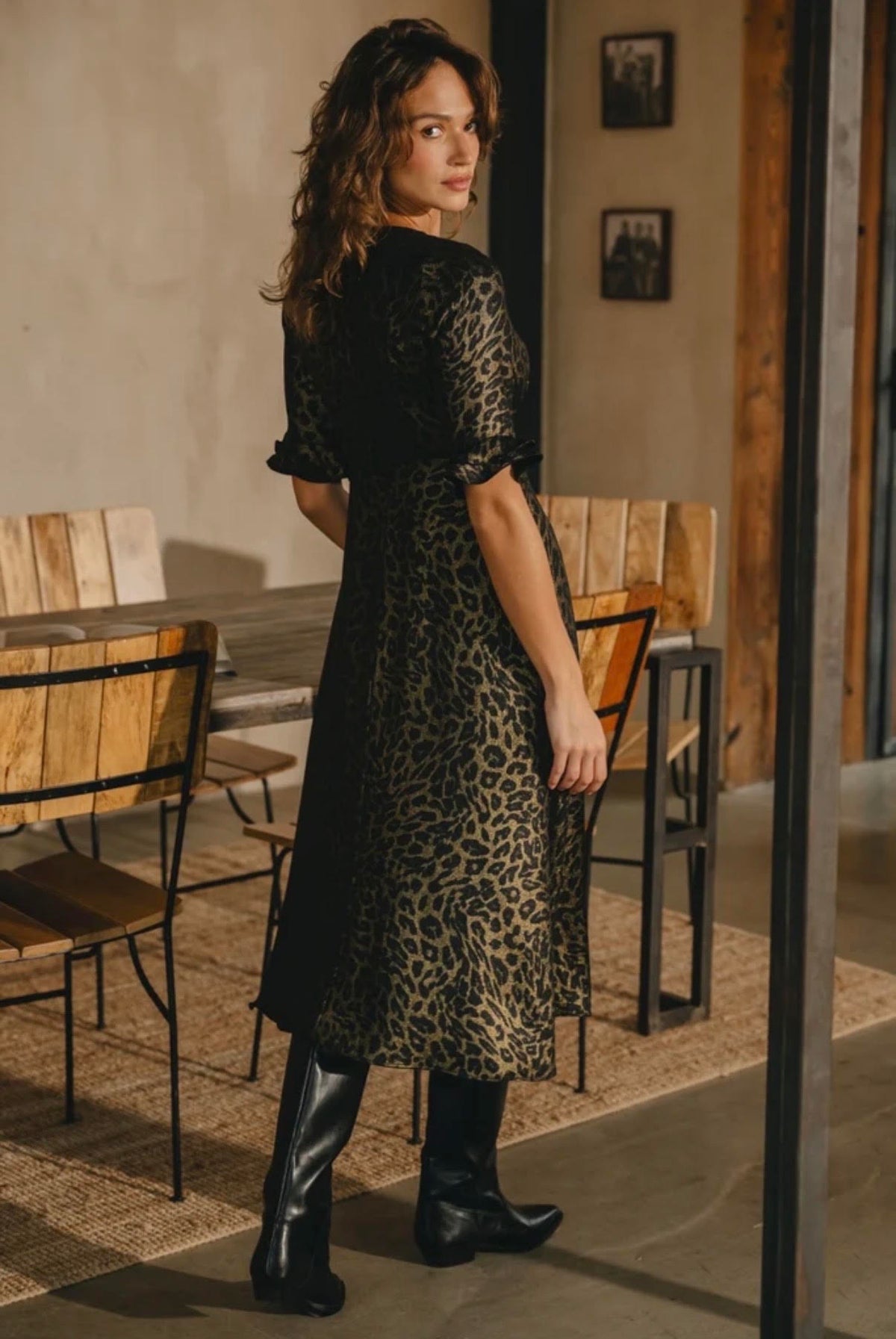 A woman wearing a black leopard-print midi dress with short sleeves and black knee-high boots, standing in a rustic indoor setting with wooden chairs and tables.