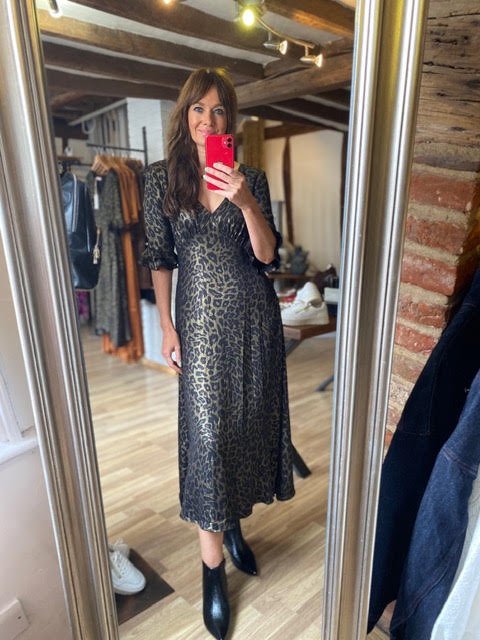 A woman takes a mirror selfie in a boutique wearing a black leopard-print midi dress with short puff sleeves and black ankle boots. The boutique has wooden floors, brick walls, and clothing racks in the background.