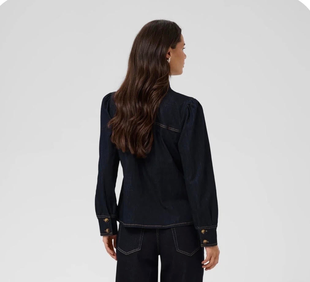 A woman with long, wavy brown hair is shown from the back wearing a dark indigo denim jacket with gold button accents on the cuffs, paired with dark jeans, against a light gray studio background.